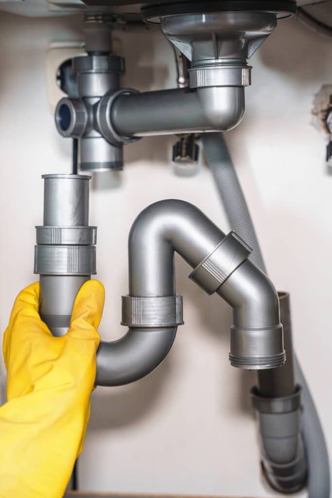 Réparation siphon évier : installation tuyauterie plastique Main gantée de jaune ajustant le siphon de canalisation en plastique gris sous un évier pour réparation de plomberie.